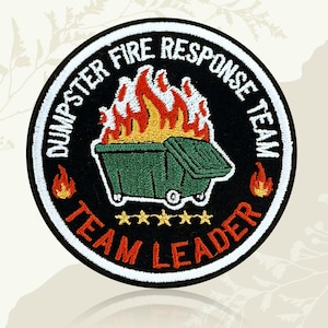 May include: A circular patch with the text "DUMPSTER FIRE RESPONSE TEAM" and "TEAM LEADER" in white lettering. The patch features a green dumpster engulfed in flames, with five gold stars below. The border is black with a white outline.
