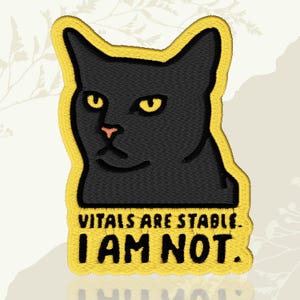 May include: Embroidered patch featuring a black cat with yellow eyes and a pink nose. The patch has a yellow border and text that reads "VITALS ARE STABLE. I AM NOT."