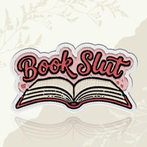 May include: Embroidered patch featuring the words "Book Slut" in red script over an open book illustration. The patch has a white border and a pink background, with a decorative floral design in the background.