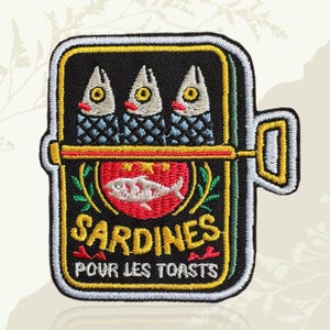 May include: A rectangular embroidered patch shaped like a sardine tin with a black background. It features three fish heads at the top, a red circle with a white fish graphic in the center, and the yellow text SARDINES POUR LES TOASTS below.