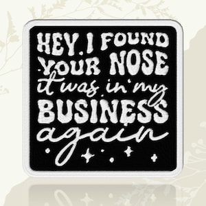 May include: A square embroidered patch with a black background and a white border. The patch features white embroidered text that reads: HEY, I FOUND YOUR NOSE IT WAS IN MY BUSINESS AGAIN, accompanied by small embroidered star and dot accents.