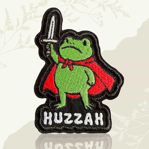 May include: Embroidered patch featuring a green frog wearing a red cape and holding a sword. The patch has a black border and the word "Huzzah" in white lettering at the bottom. The frog has a serious expression.