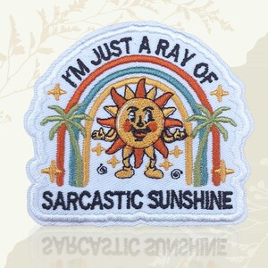 May include: Embroidered patch with the text "I'M JUST A RAY OF SARCASTIC SUNSHINE". The patch features a smiling sun character with a rainbow, palm trees, and stars in shades of orange, yellow, green, and blue.