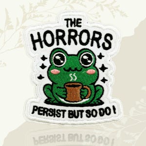 May include: Embroidered patch with a green frog holding a brown mug, and the text "THE HORRORS PERSIST BUT SO DO I" in black. The patch has a white border, black star accents, and pink cheeks. A fun, quirky design.