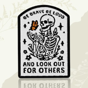 May include: Embroidered patch with a black border featuring a white skeleton holding a butterfly. The patch includes the text "BE BRAVE BE LOUD" at the top and "AND LOOK OUT FOR OTHERS" at the bottom. The design includes stars and floral elements.