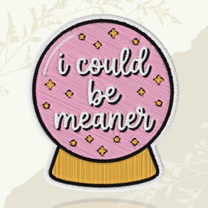 May include: Embroidered patch featuring a pink crystal ball design with the text "I could be meaner" in white script. The patch includes gold star accents and a gold base, outlined with black and white stitching.