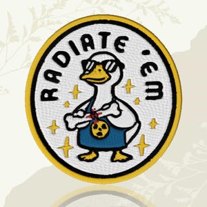 May include: Embroidered patch with a cartoon duck wearing sunglasses and a blue apron, holding a charm. The patch reads "RADIATE 'EM" in black lettering, with a yellow border and star accents.