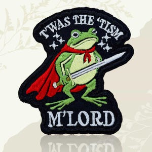 May include: Embroidered patch featuring a green frog in a red cape and holding a sword. The patch has the text "T'WAS THE 'TISM M'LORD" in white lettering on a black background. The frog has a red scarf.