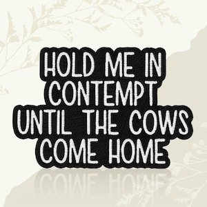 May include: Black embroidered patch with the phrase "HOLD ME IN CONTEMPT UNTIL THE COWS COME HOME" in white lettering. The patch has a black border and a textured appearance, suitable for sewing or ironing onto fabric.