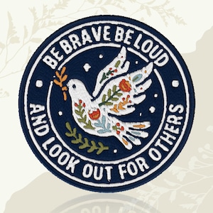 May include: Circular embroidered patch featuring a white dove with floral details in orange, green, and blue, carrying an olive branch. The navy blue patch has white text that reads "BE BRAVE BE LOUD AND LOOK OUT FOR OTHERS".