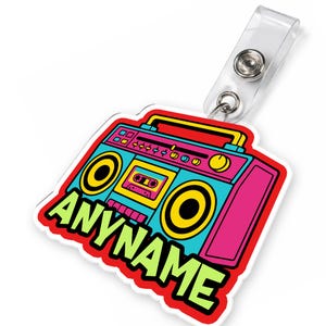 May include: A colorful, retro-style boombox badge with the text "ANYNAME" in green. The boombox design features pink, teal, yellow, and red accents. It includes a clear plastic badge holder with a metal snap.