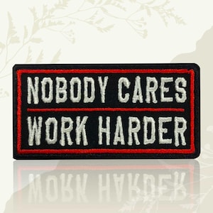 May include: A rectangular black patch with a red border and white embroidered text. The text reads "NOBODY CARES" above "WORK HARDER". The patch is likely for clothing or accessories.