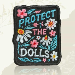 May include: Black embroidered patch featuring the text "PROTECT THE DOLLS" in white, pink, and blue. The patch is adorned with embroidered flowers and leaves in pink, white, and blue.