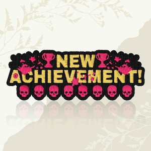 May include: Embroidered patch featuring the text "NEW ACHIEVEMENT!" in gold thread on a black base. Pink skulls and trophy icons are incorporated into the design. The patch has a decorative border and a reflective surface.