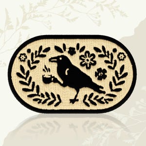 May include: An oval-shaped embroidered patch featuring a black crow holding a cup, surrounded by floral and leaf designs. The patch has a beige background and a black border, creating a decorative and nature-inspired design.