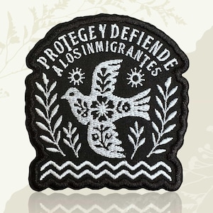 May include: A black and white embroidered patch with the Spanish text "PROTEGE Y DEFIENDE A LOS INMIGRANTES". The patch features a white dove with floral details, suns, and leafy branches. A wavy line borders the bottom.