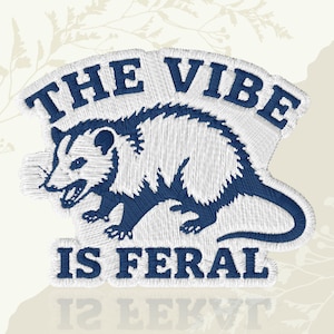 May include: Embroidered patch featuring a possum in white and navy blue. The text "THE VIBE IS FERAL" is arched above the possum and below it. The patch has a textured, stitched appearance, suitable for application on clothing or accessories.