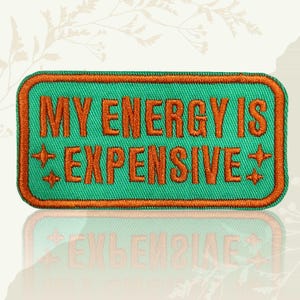 May include: A rectangular patch with the phrase "MY ENERGY IS EXPENSIVE" embroidered in orange thread on a teal background. The patch has an orange border and star-like accents. The patch is designed to be ironed on or sewn onto clothing or accessories.