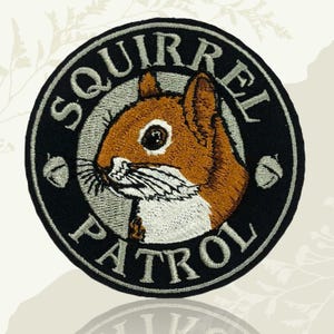 May include: Embroidered patch with a brown squirrel and white details, set on a black circular background. The words "SQUIRREL PATROL" are embroidered in grey around the squirrel. Two grey acorns are on either side of the squirrel.