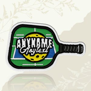 May include: Embroidered patch featuring a pickleball paddle design. The paddle has a green playing surface with white court lines, a blue area, and a yellow pickleball. The text "ANYNAME Anytext" is in white. The paddle handle is black.