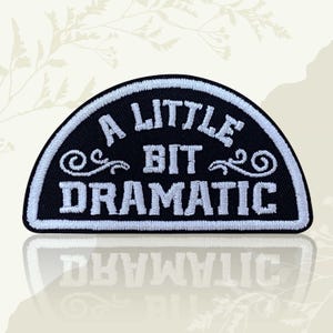 May include: A black and white embroidered patch with the words "A LITTLE BIT DRAMATIC". The text is in white, with decorative swirls. The patch is in a semi-circular shape and has a white border.