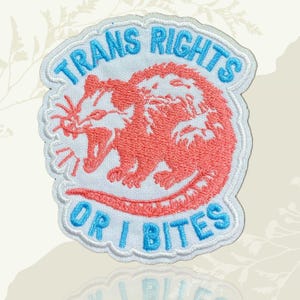 May include: Embroidered patch featuring the text "TRANS RIGHTS" above a coral-coloured opossum with an open mouth, and "OR I BITES" below. The patch has a white background and a light blue border.