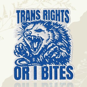 May include: Embroidered patch featuring a blue opossum with an open mouth and sharp teeth. The text "TRANS RIGHTS" is above the opossum, and "OR I BITES" is below. The design is on a cream-colored background.