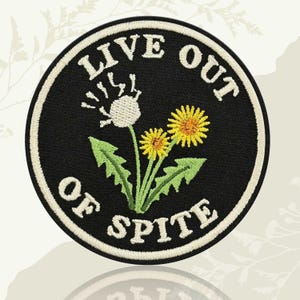May include: Embroidered patch with a black background and a white border. The patch features a dandelion design with the text "LIVE OUT OF SPITE" in white lettering. The design includes a white dandelion seed head and two yellow dandelions.