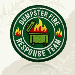 May include: A circular embroidered patch with a dark green background and a white border. The centre features a green dumpster with yellow and red flames, flanked by two small flames. The text DUMPSTER FIRE RESPONSE TEAM is stitched in white.