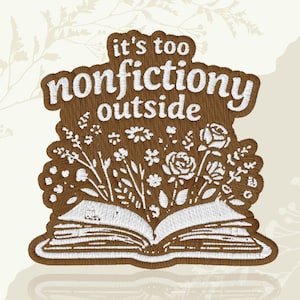 May include: Embroidered patch with the text "it's too nonfictiony outside" above an open book with floral designs. The patch is brown with white stitching, and the flowers include roses and other blooms.