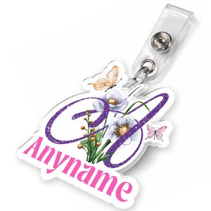 May include: A personalised name badge with a floral and butterfly design. The badge features a purple glittery initial and the name "Anymame" in pink. Includes a clear plastic strap with a metal snap.