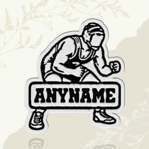 May include: Embroidered patch featuring a wrestler in a crouched position, rendered in black and white. The wrestler is wearing a singlet and headgear. Below the wrestler is a rectangular banner with the text "ANYNAME".