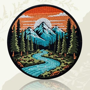 May include: A circular embroidered patch featuring a landscape scene with a winding blue river in the foreground, surrounded by green pine trees. In the background, snow-capped blue mountains rise beneath a bright orange sunset sky.