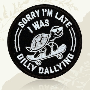 May include: Black circular patch with white embroidered text and a skateboarding turtle graphic. The text reads "SORRY I'M LATE I WAS DILLY DALLYING". The turtle wears sunglasses and rides a skateboard.