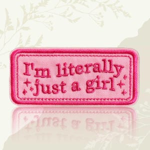 May include: A pink rectangular patch with the embroidered text "I'm literally just a girl" in a darker pink. The patch has a stitched border and decorative star-like accents.