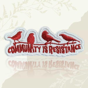 May include: Embroidered patch with four red birds on a brown branch. The text "COMMUNITY IS RESISTANCE" is embroidered in red below the birds. The patch has a white border.