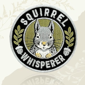 May include: Circular embroidered patch with a grey squirrel design and the words "SQUIRREL WHISPERER". The patch has a black border with white lettering, olive green accents, and a leaf and acorn design.