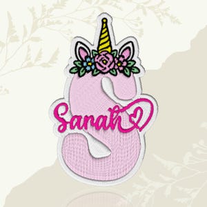 May include: Pink embroidered patch in the shape of the letter "S" with the name "Sarah" in cursive. The letter is decorated with a unicorn horn, ears, and a floral crown. The patch has a white border.