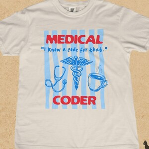 May include: An ivory short-sleeved t-shirt featuring a graphic with light blue vertical stripes. The text reads MEDICAL, I Know a code for that., and CODER in red and blue. Illustrations include a blue stethoscope, a caduceus, and a coffee mug.