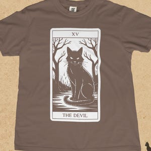 Puede incluir: Camiseta color espresso con un diseño de carta de tarot blanca que presenta una ilustración de un gato negro. La carta dice "XV" en la parte superior y "THE DEVIL" en la parte inferior. El gato está sentado en un bosque.