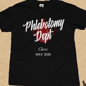 Puede incluir: Una camiseta negra de manga corta con el texto blanco Phlebotomy Dept en fuente cursiva, situado sobre un gráfico de gota de sangre roja con una línea de latido. Debajo del gráfico, el nombre Chris y el texto SINCE 2020 están impresos en blanco.
