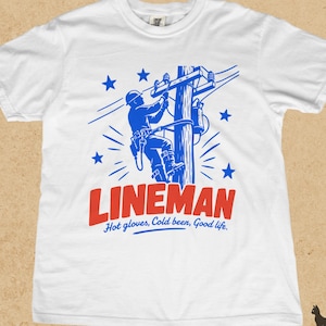 May include: A white t-shirt featuring a blue graphic of a utility worker on a power pole. The text LINEMAN appears in large red letters below the graphic, followed by the blue text Hot gloves, Cold beer, Good life. Blue stars surround the worker.