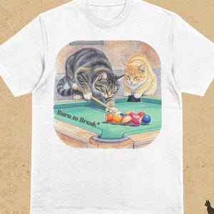 Puede incluir: Camiseta blanca con una colorida ilustración de dos gatos jugando al billar. Un gato está a punto de golpear las bolas con un taco. El texto "Born to Break" está impreso en la mesa de billar. La palabra "WHITE" está impresa en la parte superior.