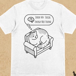 Puede incluir: Camiseta blanca con un gráfico en blanco y negro de un gato en un sofá. Una burbuja de pensamiento sobre el gato dice "ERROR 404: SOCIAL ENERGY NOT FOUND". La palabra "WHITE" está impresa en la parte superior izquierda.