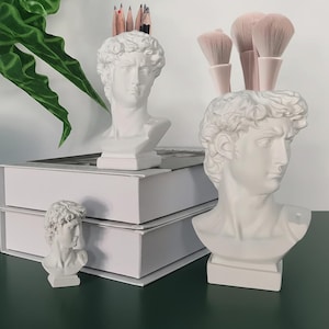 May include: White bust of David sculpture used as a pencil holder, makeup brush holder, and decorative item. The bust is on a white base and has a classic, detailed design. Pencils and makeup brushes are visible.