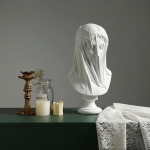 Può includere: Busto bianco di una figura velata. La scultura è su un piedistallo. Un candelabro dorato, un barattolo di vetro con una candela e una piccola candela sono su una superficie verde scuro.