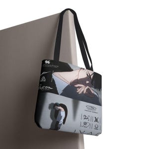 May include: Black tote bag with a grayscale anime illustration. The bag has black handles and a white background with graphic elements and text. The bag is hanging on a neutral-coloured wall.