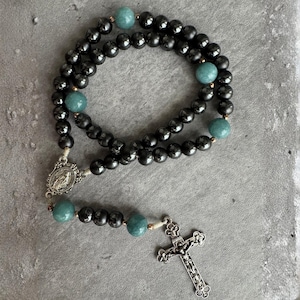May include: A rosary featuring smooth, round, dark grey hematite beads interspersed with larger, round, light green jade-like beads. The piece includes a silver-toned metal centrepiece depicting a religious figure and a matching ornate crucifix.