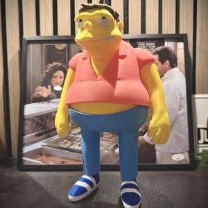 May include: A 3D-printed figure of a cartoon character with yellow skin, a red shirt, blue trousers, and blue and white striped shoes. The figure is in front of a framed picture. The figure is a character from The Simpsons animated television show.