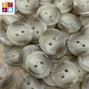 Puede incluir: Un conjunto de botones redondos de color blanco roto con efecto veteado. Cada botón tiene dos agujeros para coser. Los botones son de color neutro, adecuados para diversos proyectos de costura.